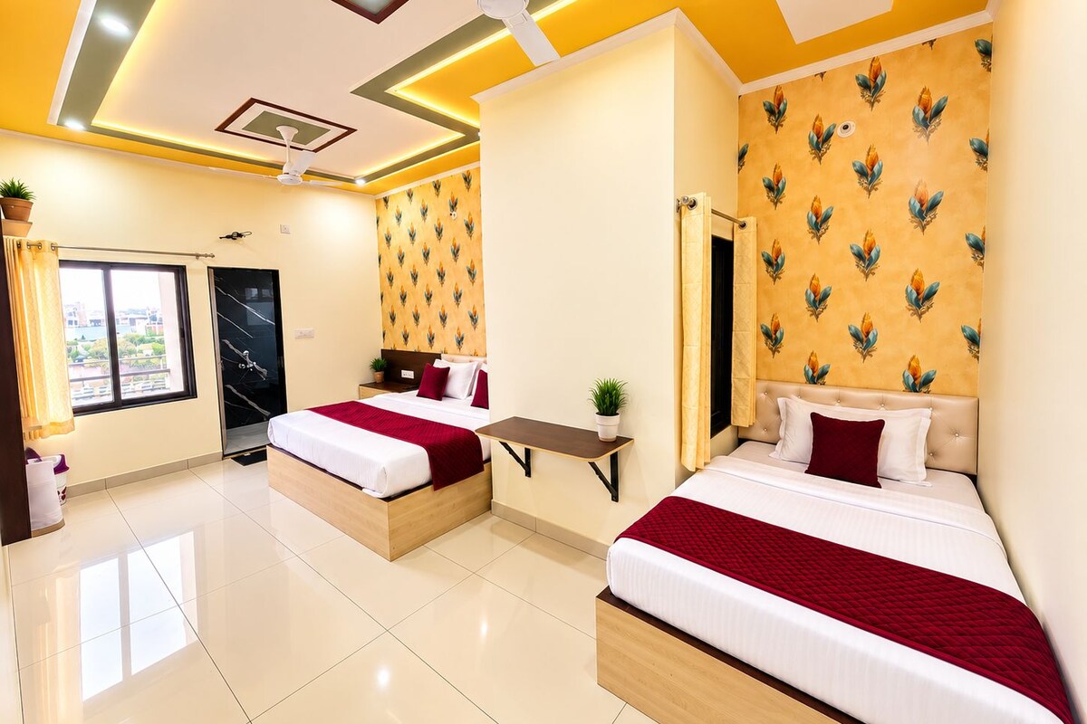 A hotel room with two beds, a small table, a ceiling fan, and a window with curtains.