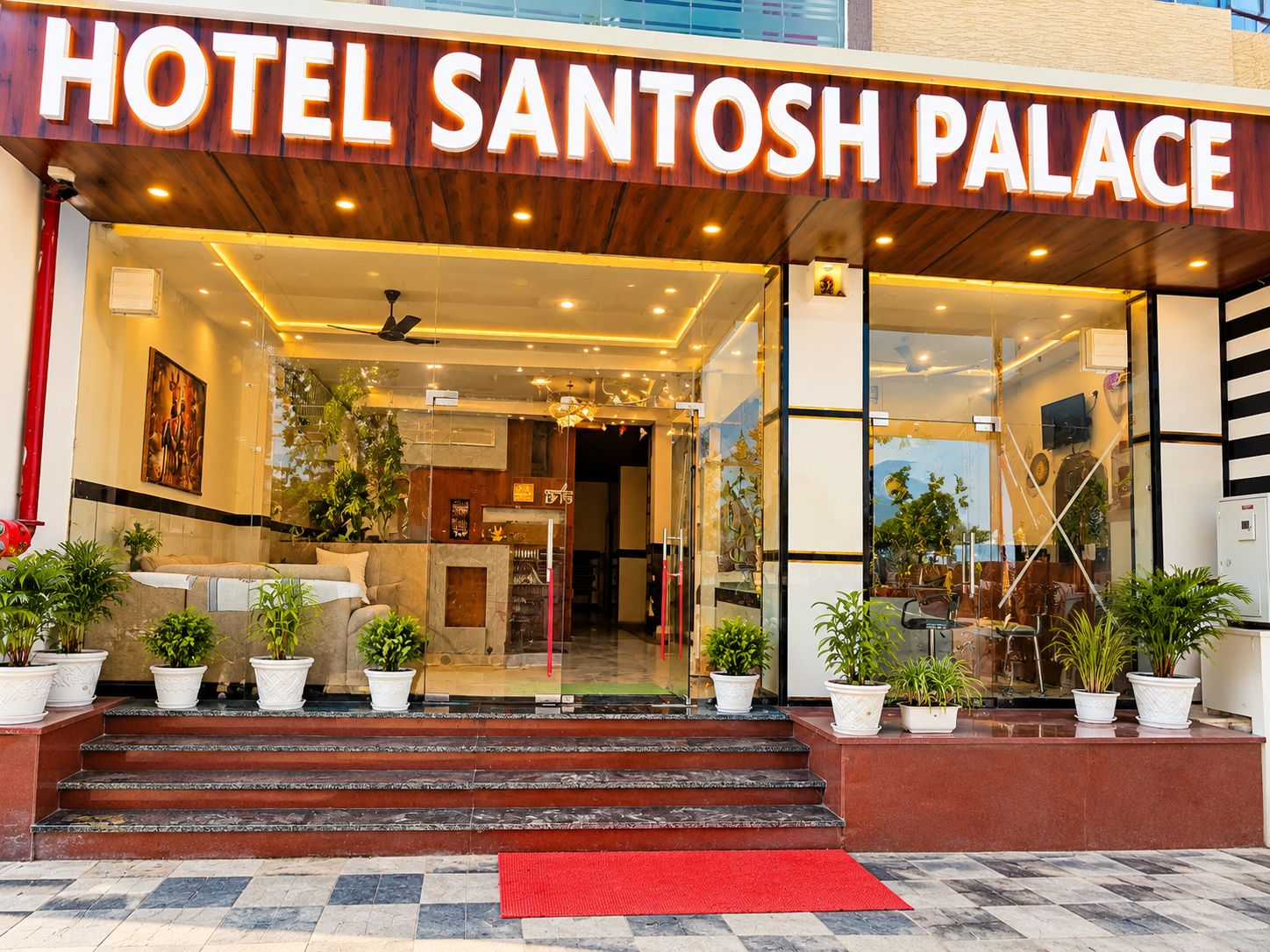 La entrada del Hotel Santosh Palace con un diseño moderno, que presenta un frente de madera, puertas de cristal y plantas en macetas.
