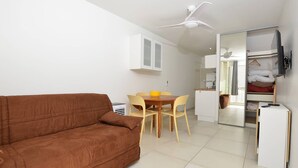 A compact living space with a brown sofa, a dining table with chairs, a kitchenette, a ceiling fan, and a TV mounted on the wall. - Studio 150m from the beach - 2 guests (Le Grau-du-Roi)