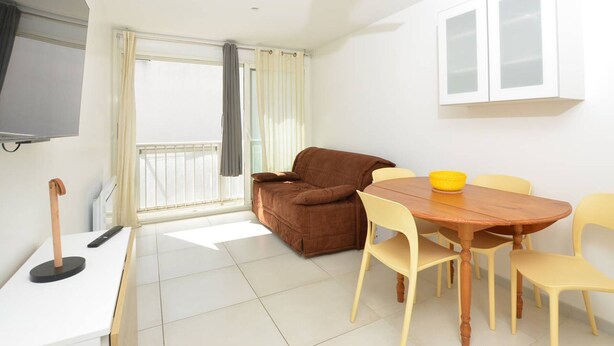 A compact living space with a sofa, dining table, chairs, a ceiling fan, a TV, and a window with curtains. - Studio 150m from the beach - 2 guests (Le Grau-du-Roi)