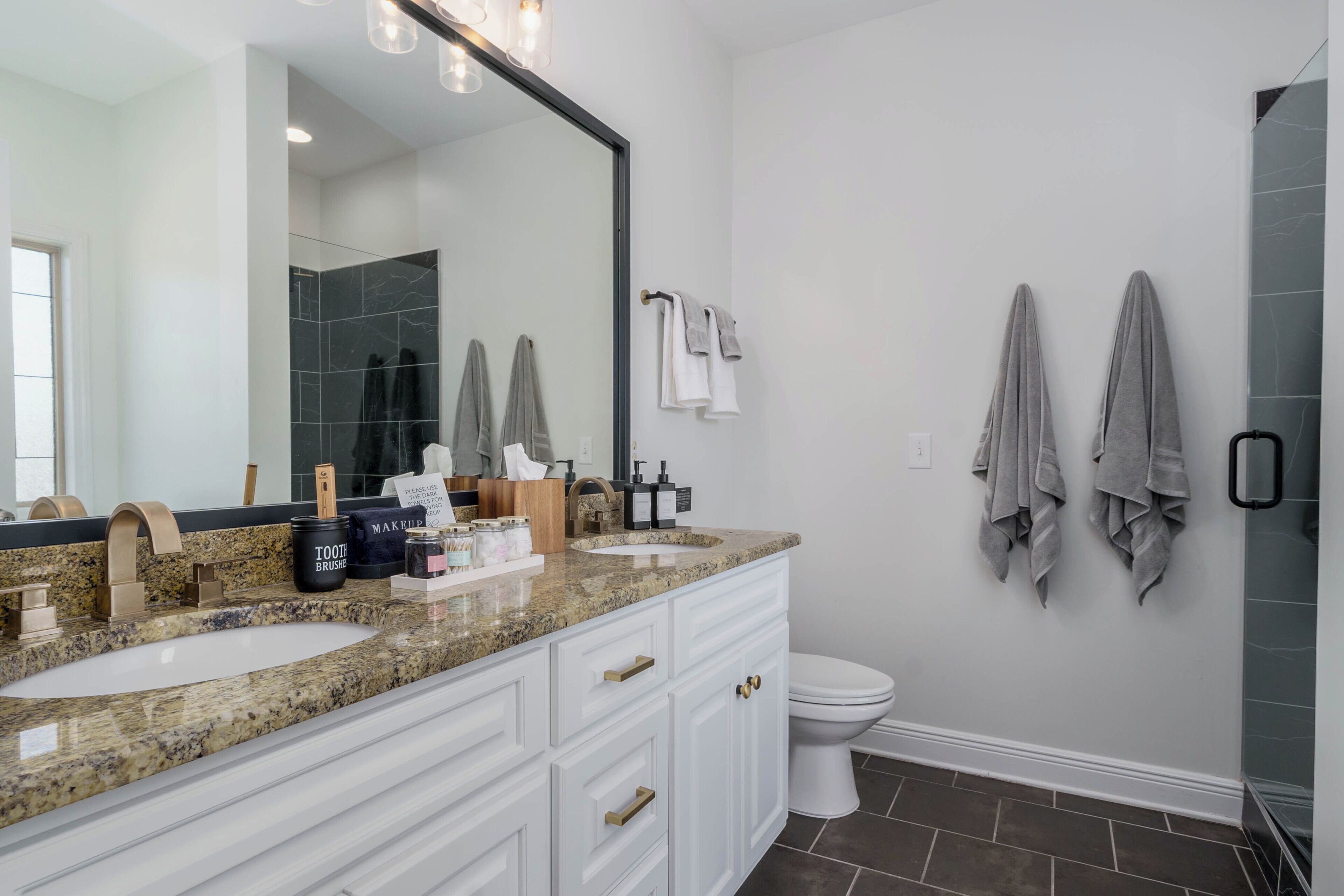A bathroom with a large mirror, two sinks, a toilet, and towels on the wall.