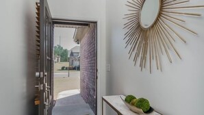 Ein Flur mit einer zur Außenseite führenden Tür, einem an der Wand befestigten Spiegel und einem kleinen Tisch mit einer Schale Grünpflanzen.