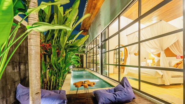Zona de piscina con terraza de madera, sillas de descanso y vista a una habitación con cama y cortinas.