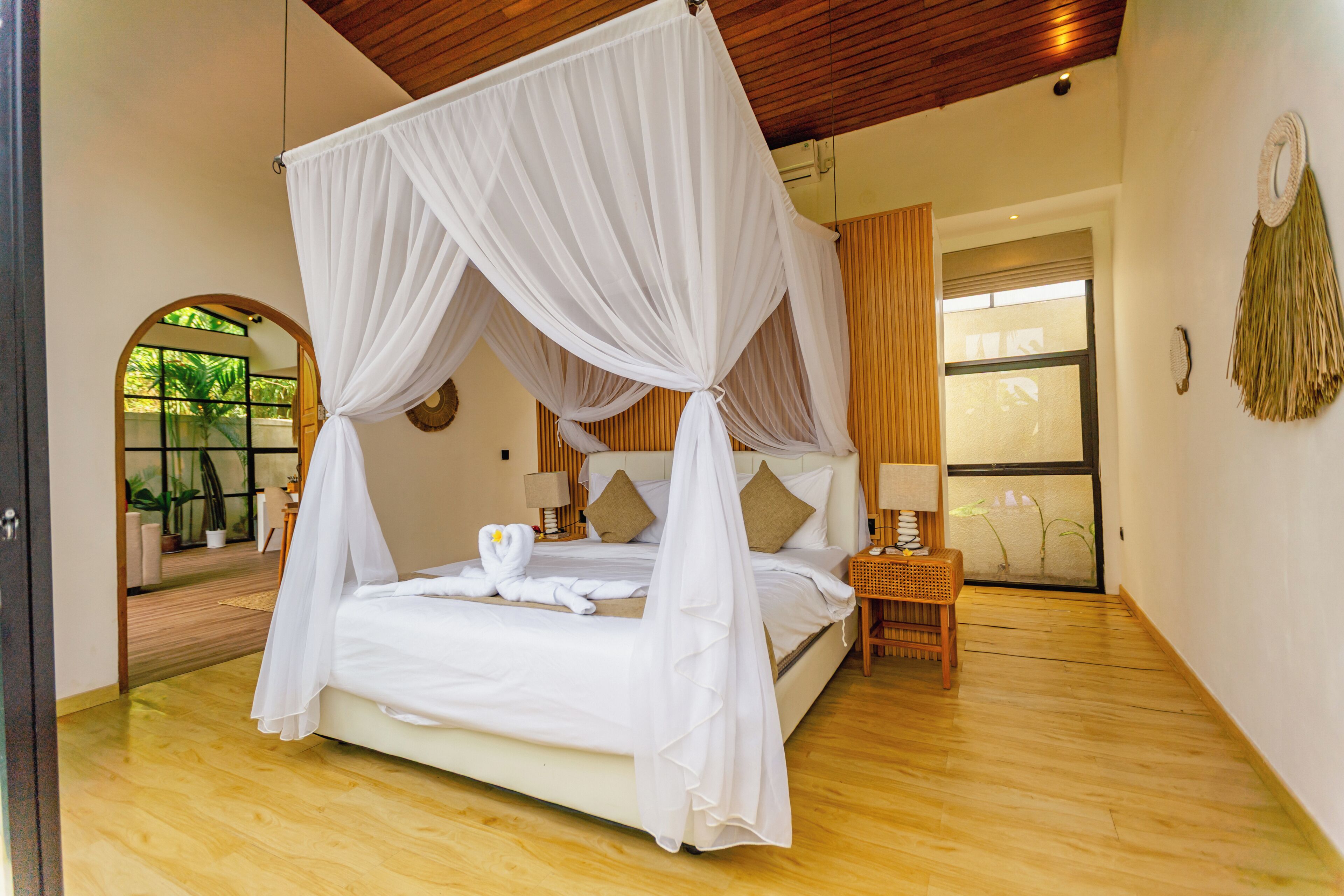 Una cama con dosel sobre cuatro columnas y dosel blanco, suelo de madera, mesita de noche con lámpara y vistas al exterior a través de una ventana.