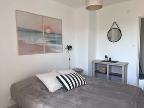 Un dormitorio con cama, lámpara de noche, una mesita de noche con espejo y un cuadro colgado en la pared.