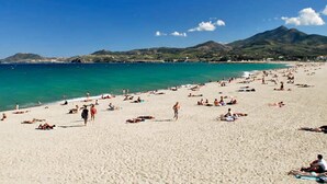 Ein Strand mit Menschen, die sonnenbaden und schwimmen, klares blaues Wasser und Bergen im Hintergrund.