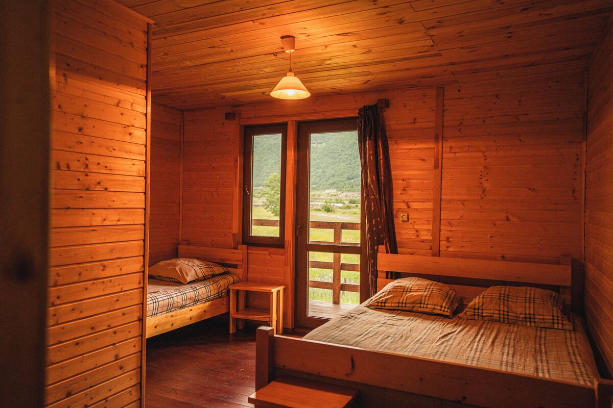 A wooden cabin room with two beds, a small table, and a window with a view of greenery.