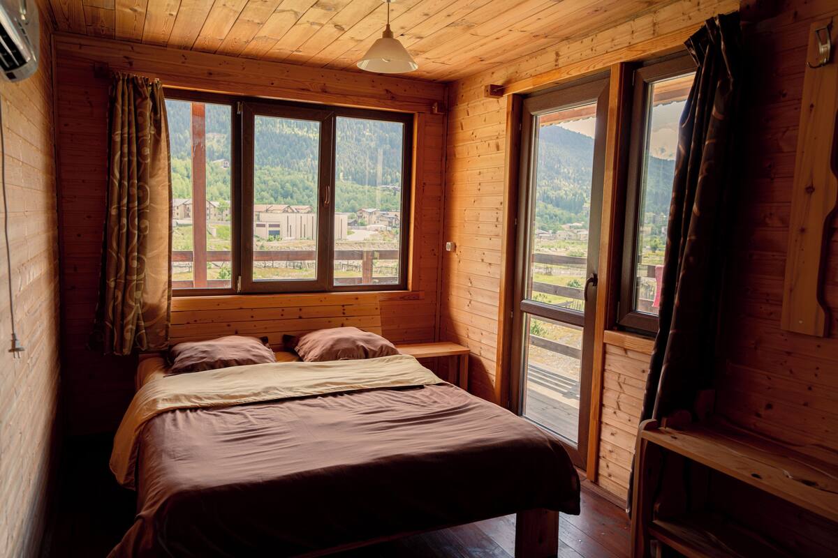 A wooden cabin bedroom with a bed, two windows, and a view of a mountainous landscape.