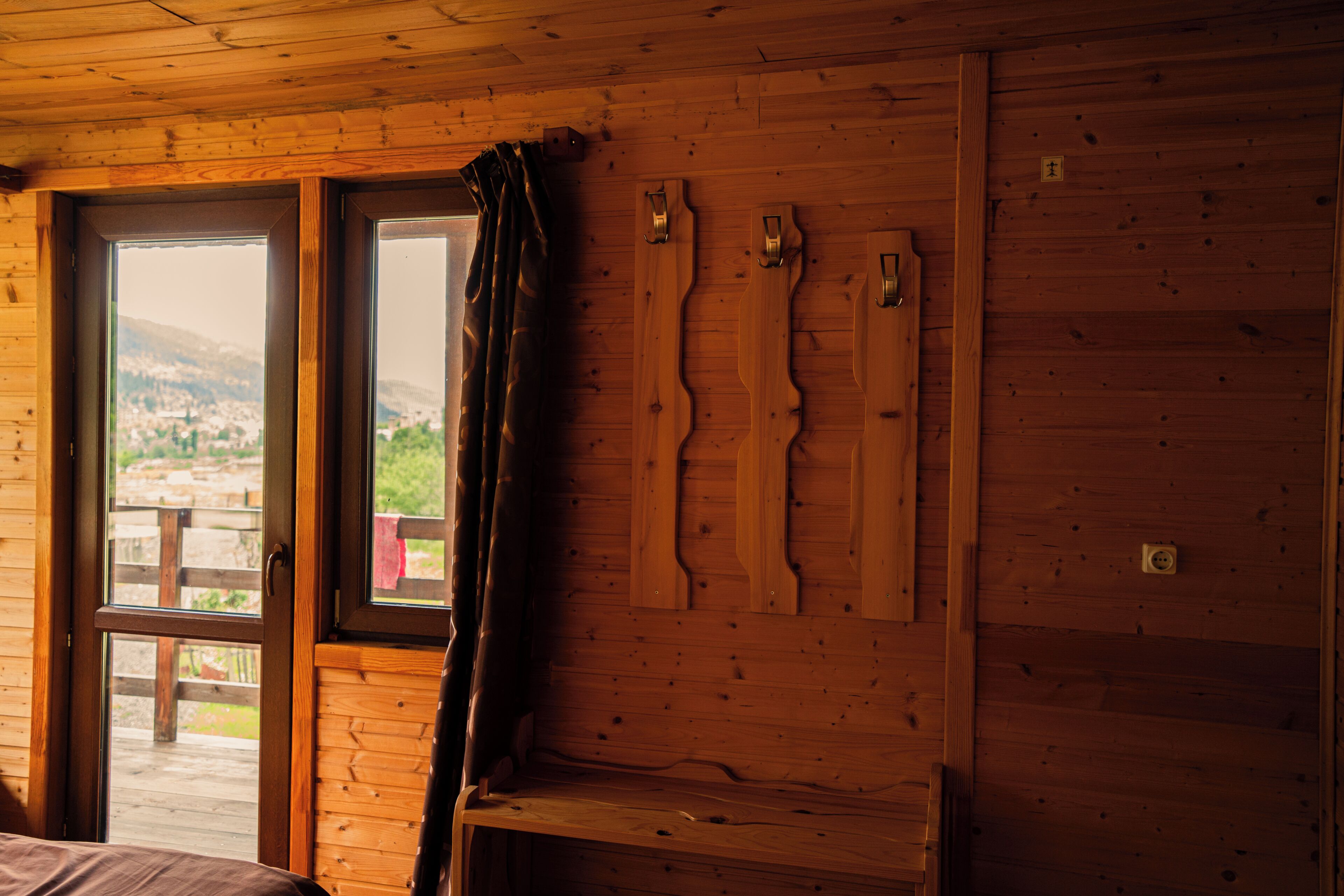 A wooden cabin interior with a large window, wooden wall-mounted hooks, and a bench.