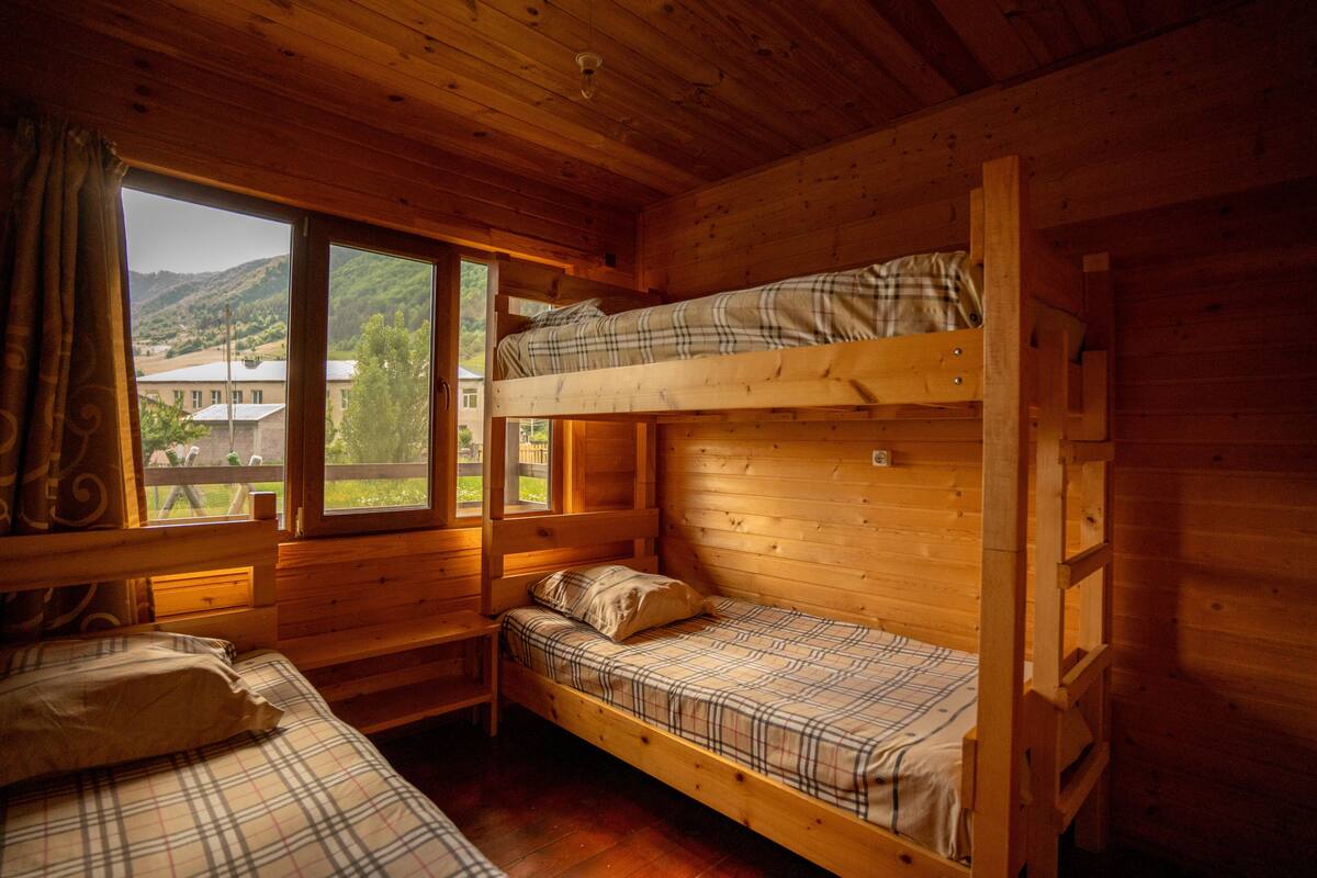 A cabin with two bunk beds, a window with curtains, and a view of a house and hills.