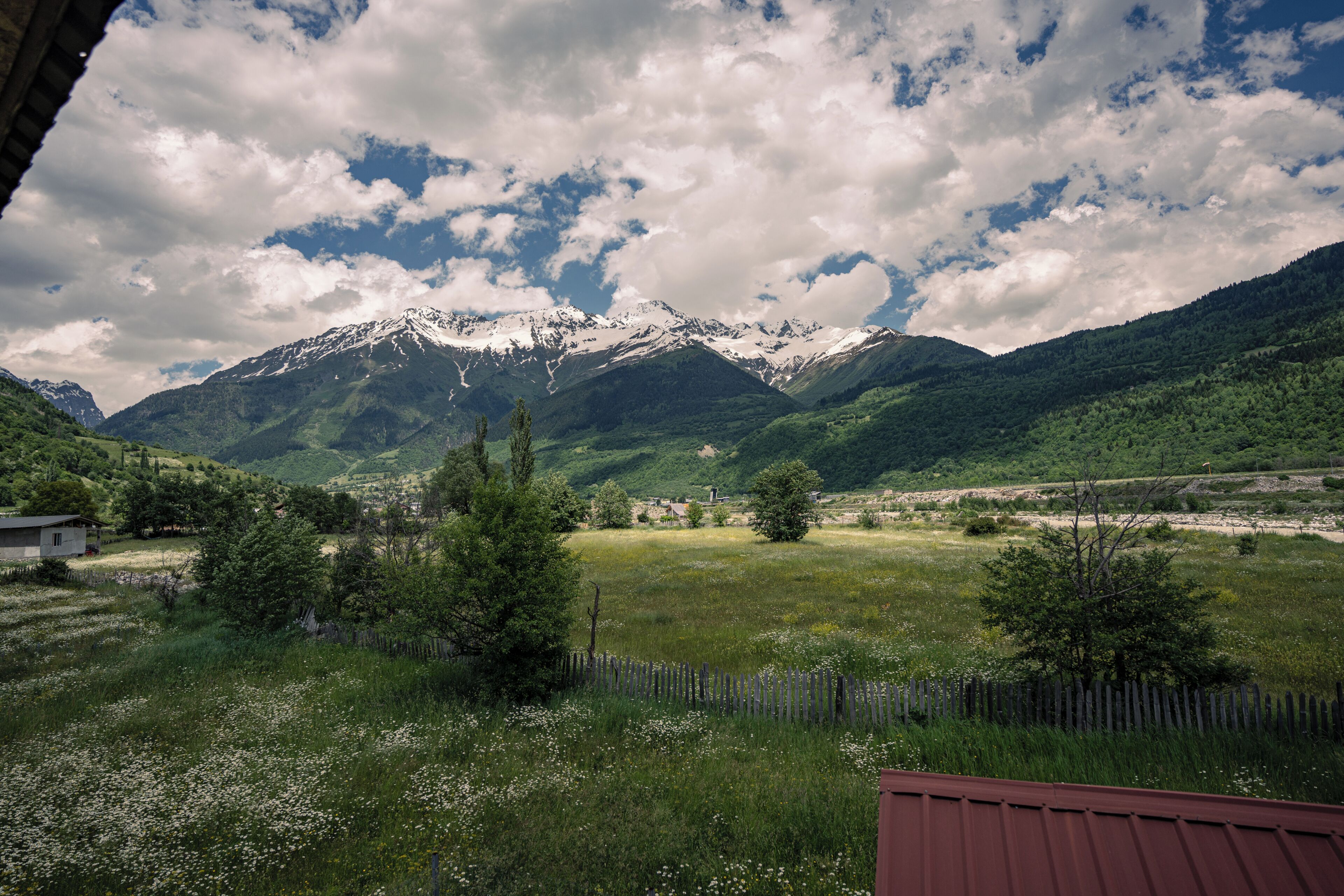 A mountainous landscape with snow-capped peaks, a grassy field, and a wooden fence.