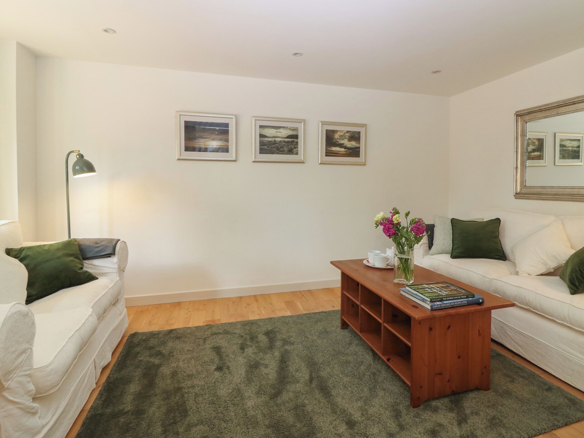 Ein Wohnzimmer mit einem weißen Sofa, einem Holztisch mit Büchern, einem grünen Teppich und drei gerahmten Bildern an der Wand.