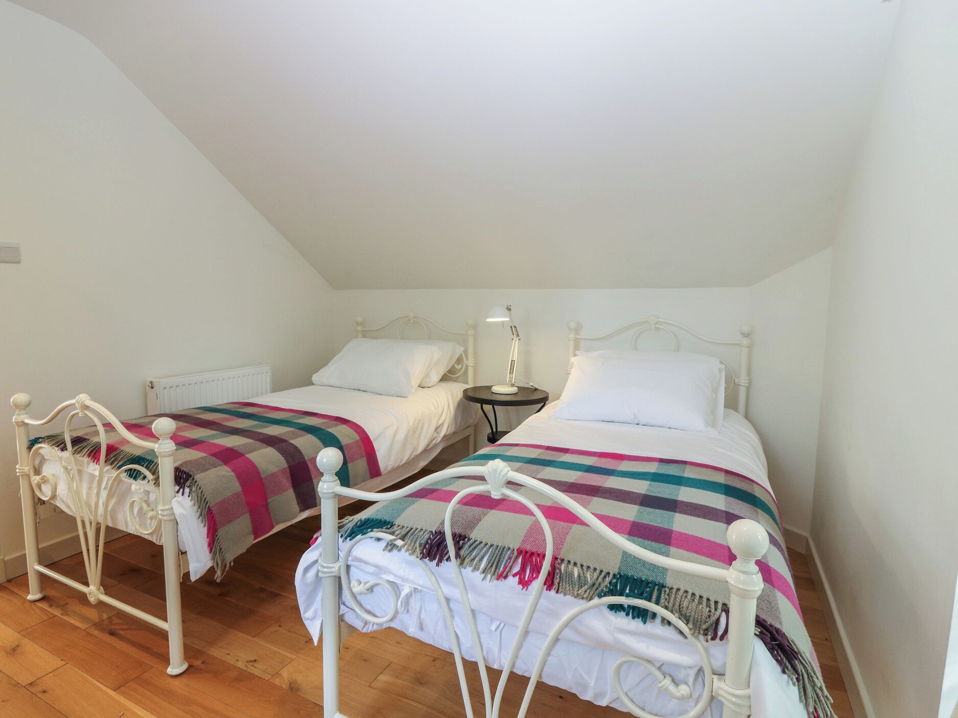 Two twin beds with white frames and colorful blankets, a small bedside table with a lamp, and a radiator on the wall.