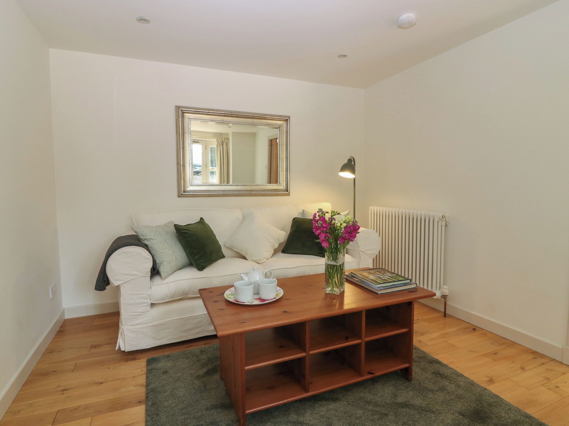 A living room with a white sofa, a wooden coffee table, a mirror, a radiator, and a green rug.