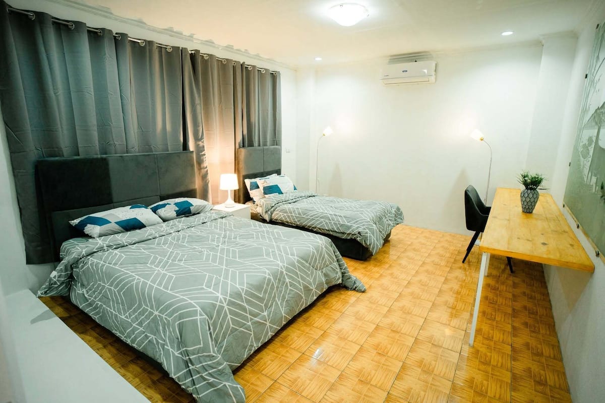 Una habitación con dos camas, una mesa de madera, una silla, una lámpara y un equipo de aire acondicionado.
