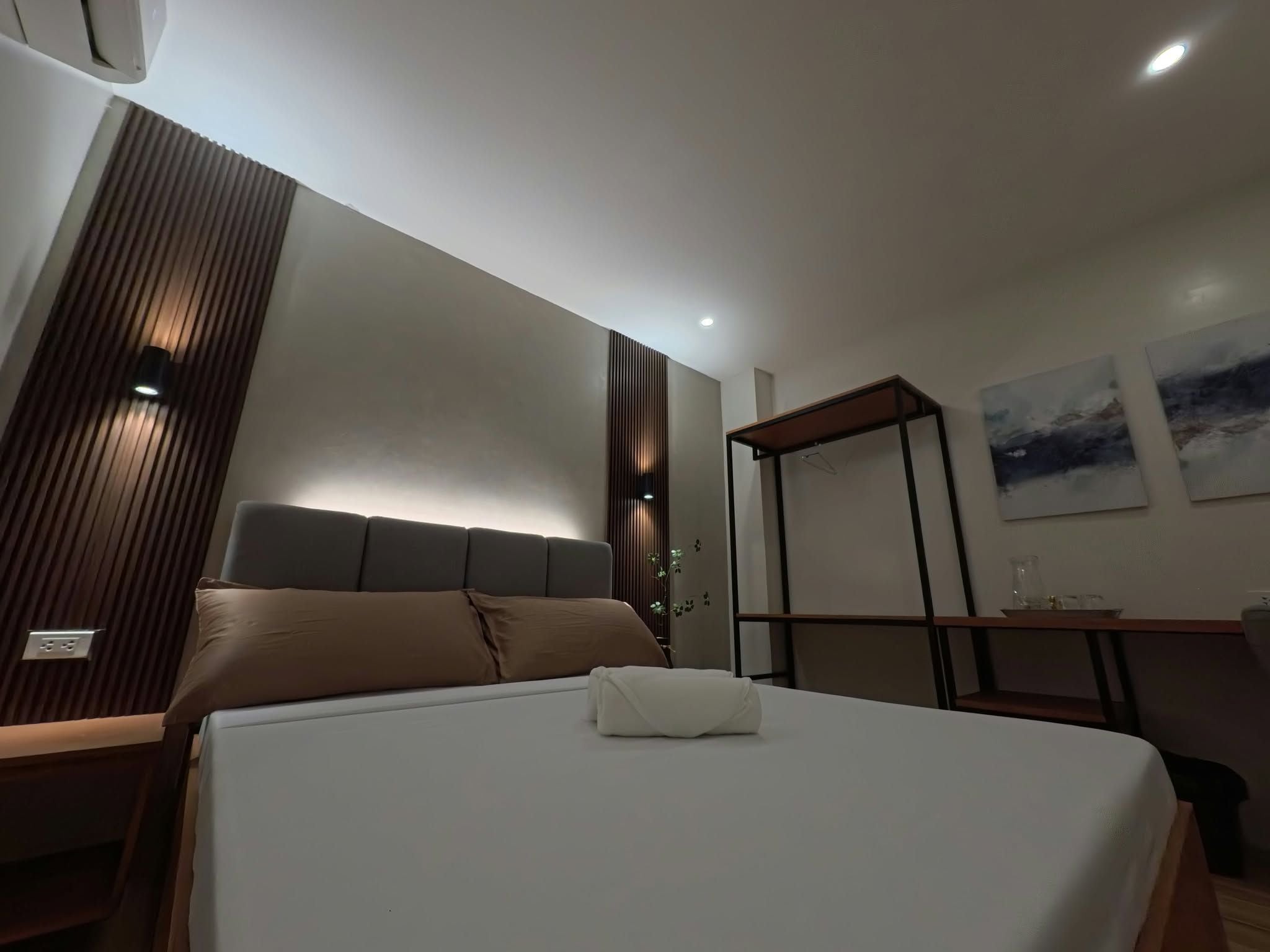 A modern hotel room with a large bed, a bedside table, a wardrobe, and a small desk with a chair.