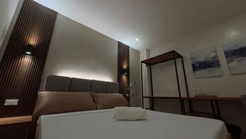 A modern hotel room with a large bed, a bedside table, a wardrobe, and a small desk with a chair.