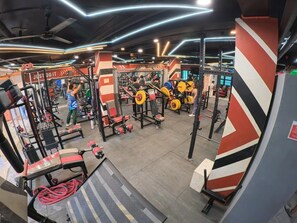 A gym with various weightlifting equipment and machines, a red and white striped wall, and a ceiling with linear light fixtures.