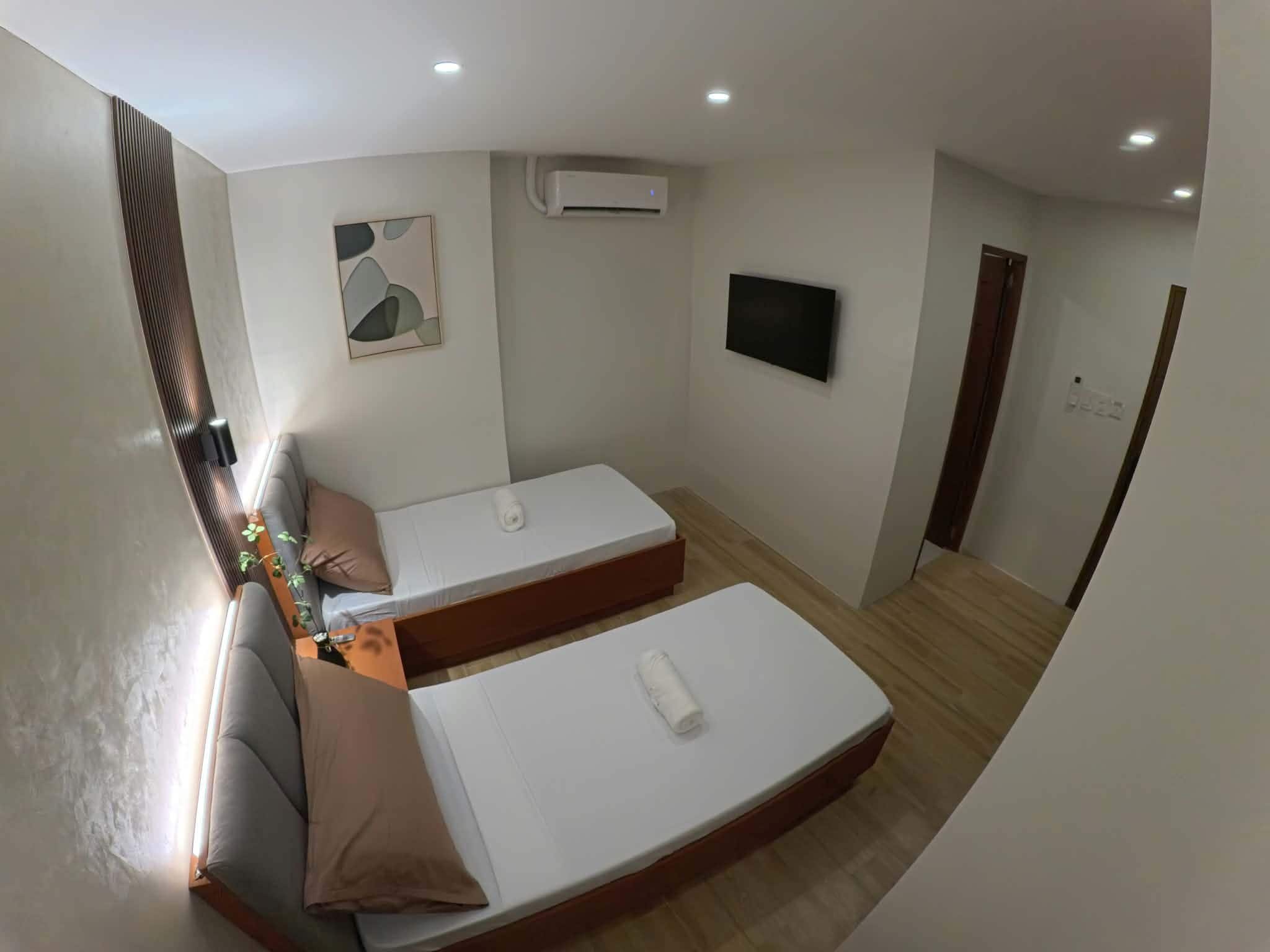 Een compacte kamer met twee eenpersoonsbedden, een klein tafeltje met een plant, een wandtelevisie en een airconditioning.