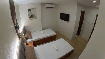 A compact room with two single beds, a small table with a plant, a wall-mounted TV, and an air conditioning unit.