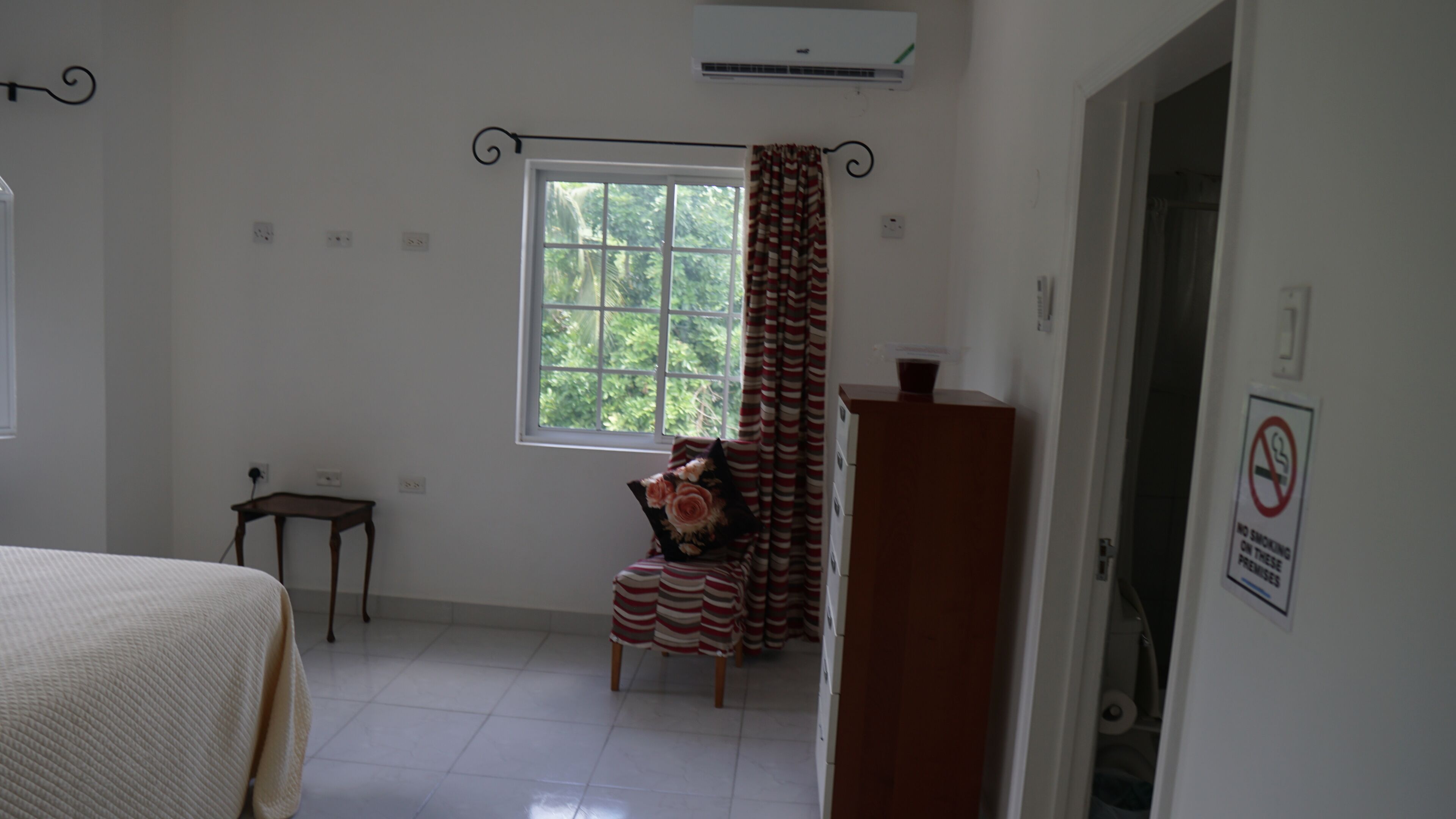 A room with a bed, a small table, a chair with a floral cushion, a window with curtains, and a no-smoking sign.