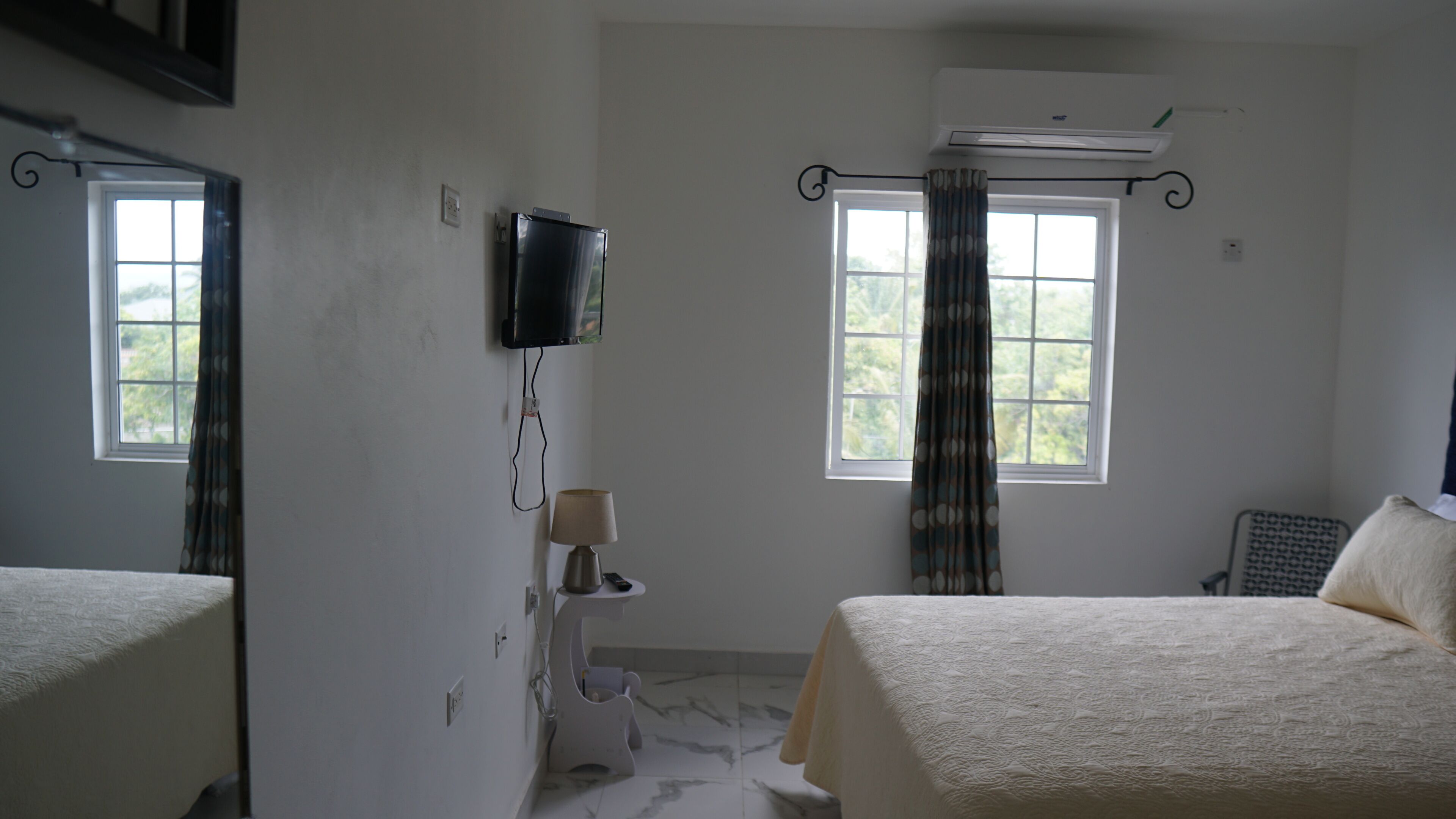 A hotel room with two beds, a television mounted on the wall, an air conditioning unit, a window with curtains, and a small table with a lamp.
