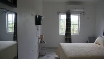 A hotel room with two beds, a television mounted on the wall, an air conditioning unit, a window with curtains, and a small table with a lamp.