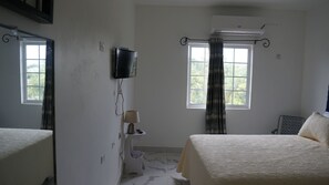 Ein Hotelzimmer mit zwei Betten, einem an der Wand befestigten Fernseher, einer Klimaanlage, einem Fenster mit Vorhängen und einem kleinen Tisch mit einer Lampe.