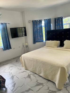 A bedroom with a bed, a flat-screen TV, a window with curtains, and a marble floor.
