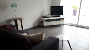 A modern living room with a dark sofa, a small round table, a TV on a white entertainment unit, and a potted plant by the window. - House on the Arcachon Bay with Air Conditioning (Gujan-Mestras)
