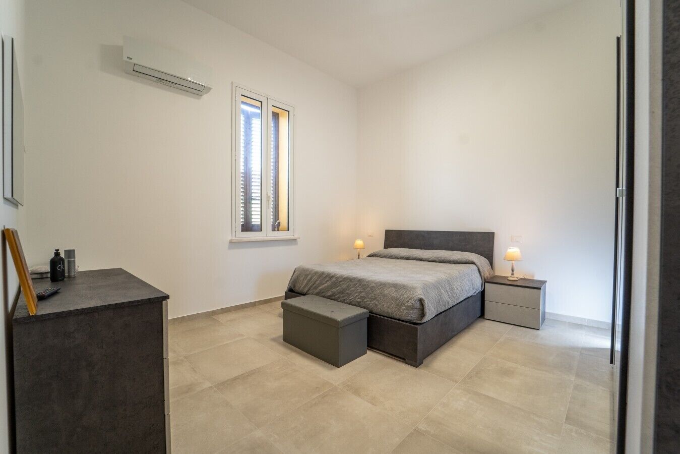 A bedroom with a bed, bedside tables, a desk, a window, and an air conditioning unit.