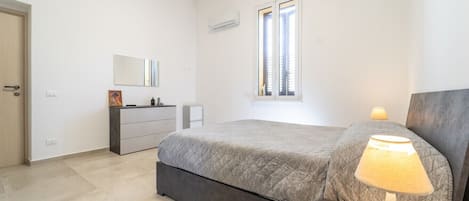 A bedroom with a bed, bedside table, lamp, dresser, mirror, and window.