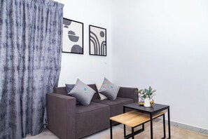 A modern living room with a grey sofa, two cushions, a small wooden table, and abstract wall art. - Bloomfields 1BR in Oasis Masdar  (Abu Dhabi)