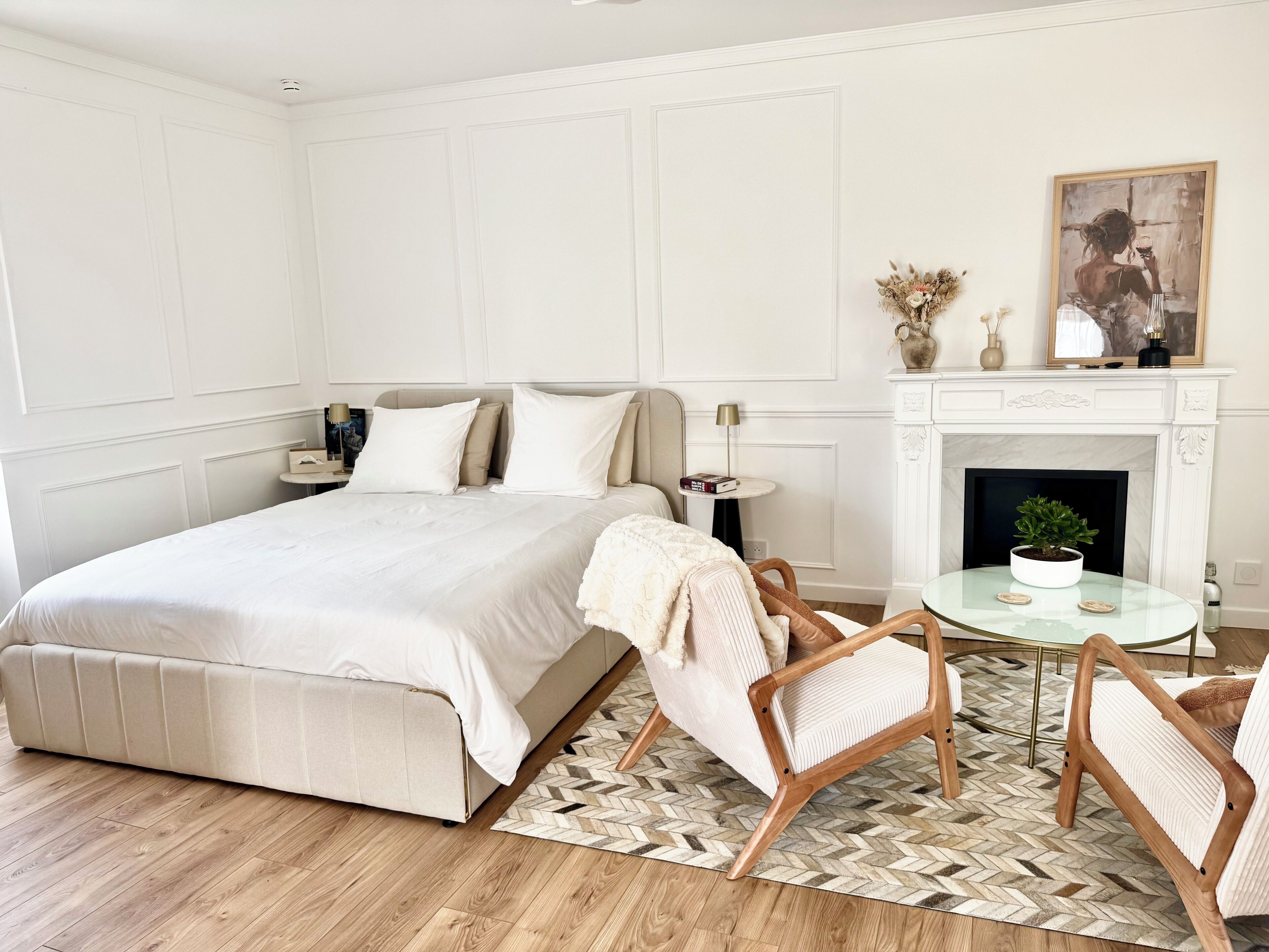 A bedroom with a large bed, a fireplace, a small table with a plant, and two chairs.