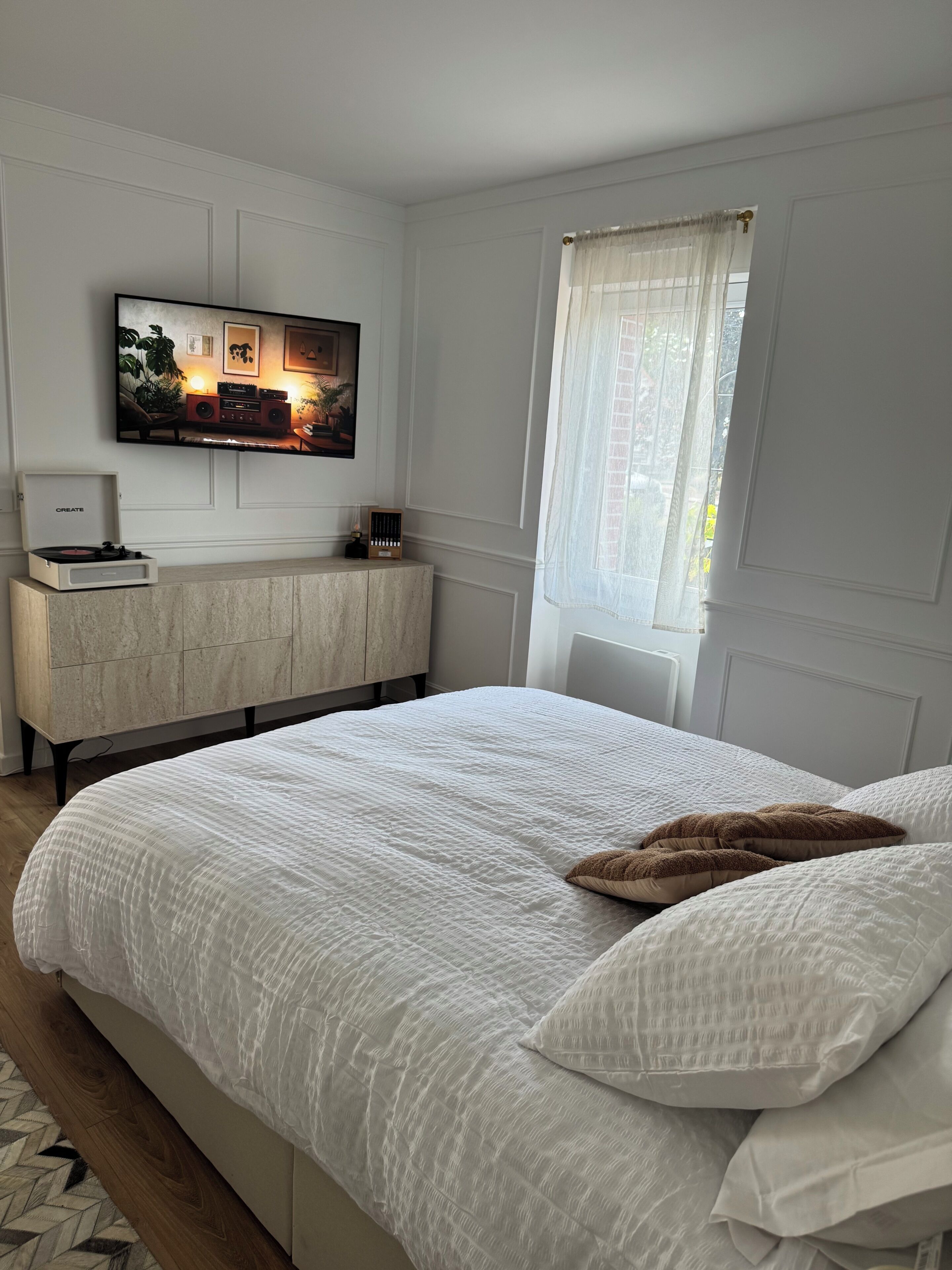 Une chambre à coucher avec un grand lit, un téléviseur fixé au mur, une armoire en bois et une fenêtre ornée de rideaux légers.