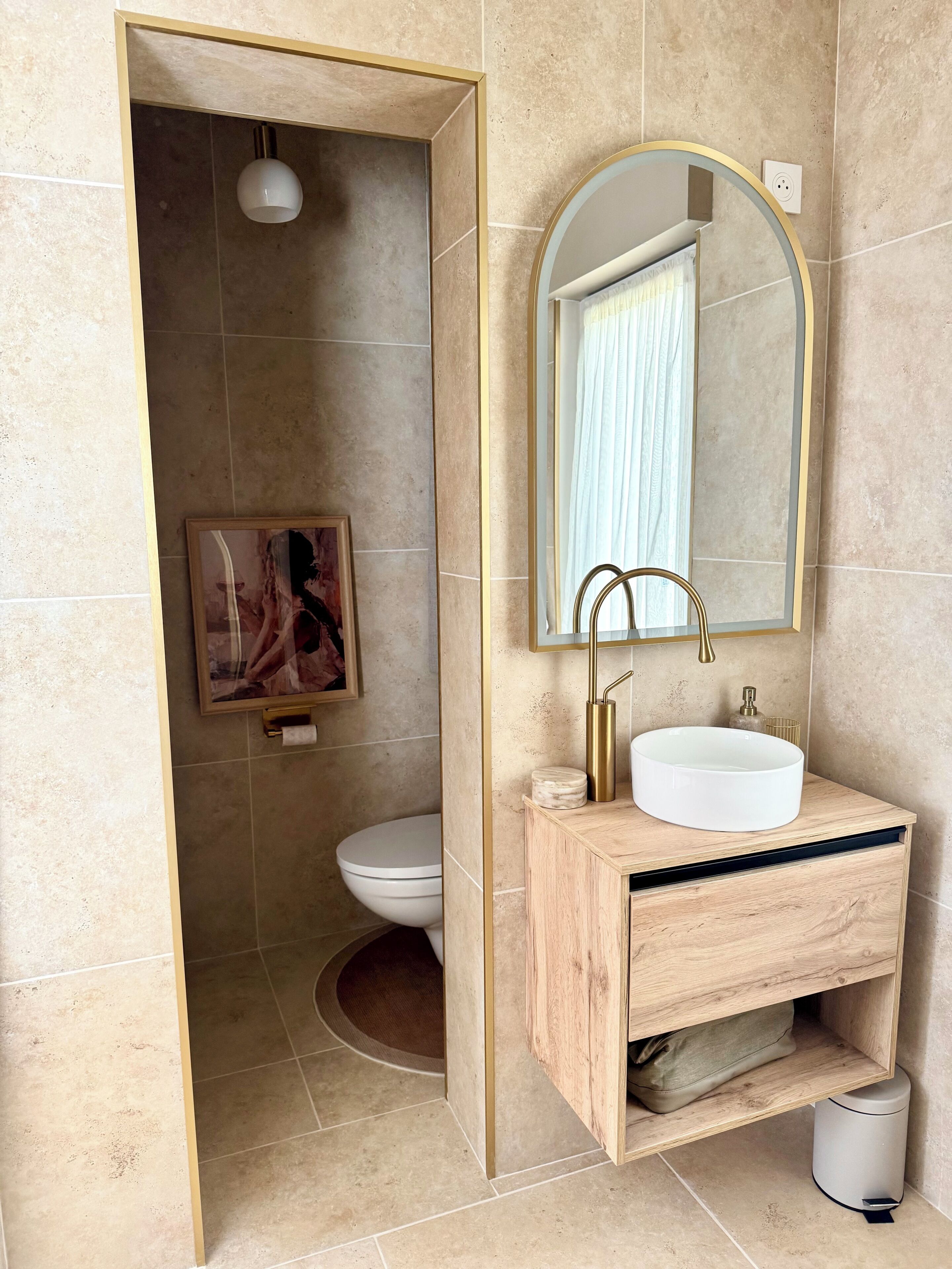 Un bagno con un mobile contenitore in legno, un lavandino bianco, uno specchio con cornice ad arco, un water e una zona doccia.
