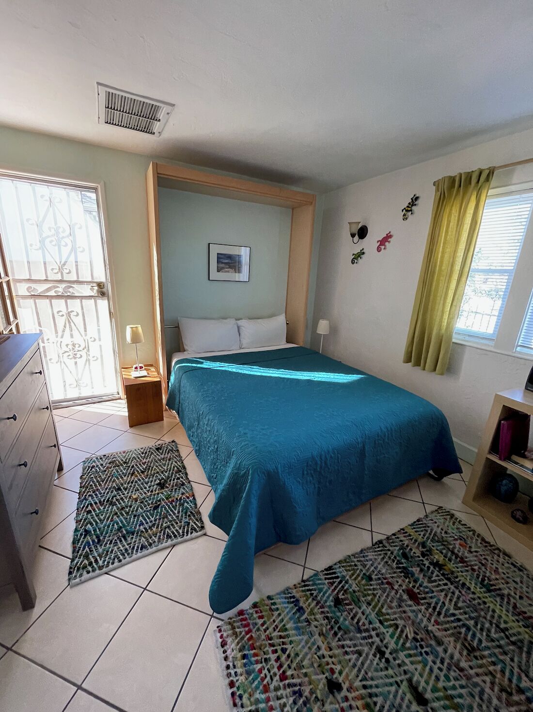 A bedroom with a bed, a dresser, a nightstand, a window with curtains, and a wall-mounted air vent.