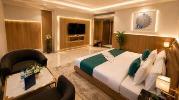 A modern hotel room with a large bed, a seating area with a sofa and armchair, a flat-screen TV, and a wall-mounted artwork.