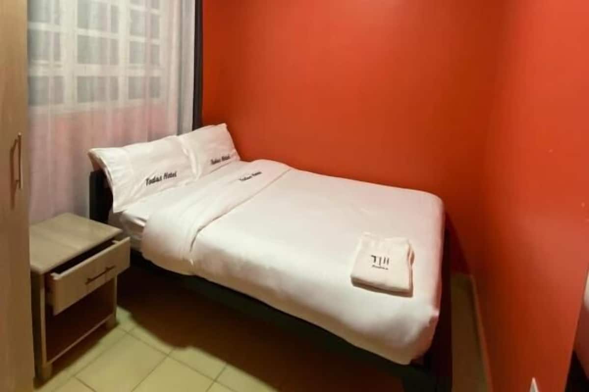 A single-bed room with a wooden wardrobe, a small bedside table, and a window with a curtain.