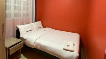 A single-bed room with a wooden wardrobe, a small bedside table, and a window with a curtain.