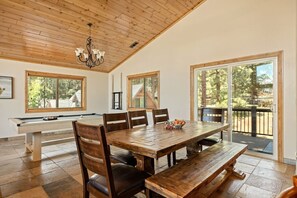 A dining area with a wooden table and chairs, a chandelier, and large windows with a view of trees. - Snowboard Shores| Game Room | Hot Tub | Lake View (Big Bear Lake)