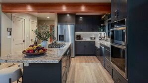 A modern kitchen with dark cabinetry, stainless steel appliances, a marble countertop, and a central island with fruit. - Pine & Paw | Hot Tub, Sauna, Games, Views, Piano (Big Bear Lake)