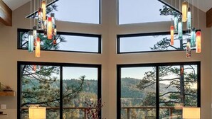A spacious room with a large window, a sofa, a table, and hanging lights. - Pine & Paw | Hot Tub, Sauna, Games, Views, Piano (Big Bear Lake)