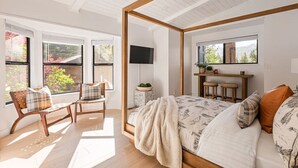 Quarto com cama de dossel, uma escrivaninha com dois banquinhos, uma TV fixada na parede e uma janela com vista para árvores.