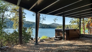 Uma área externa coberta com uma banheira de hidromassagem, assentos e vista para um lago e montanhas.