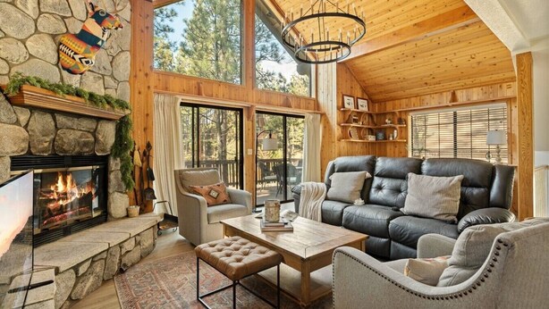Interior acogedor de una cabaña con chimenea de piedra, sofá de cuero y un ventanal con vistas a árboles.