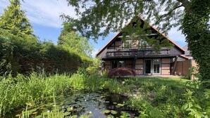 A modern two-story house with a large garden, a pond with lily pads, and a wooden deck.