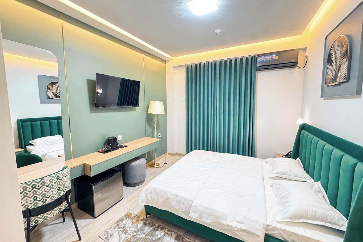 Quarto de hotel com cama, escrivaninha com cadeira, televisão, luminária e janela com cortinas.