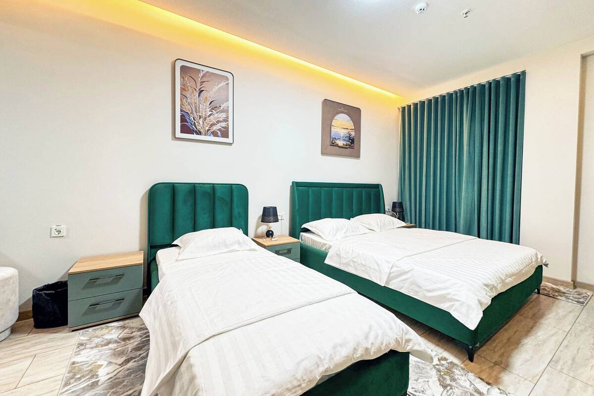 Quarto de hotel com duas camas, uma mesinha de cabeceira, uma luminária, janela com cortinas e um quadro na parede.