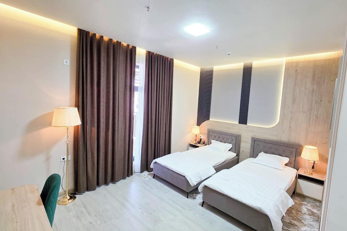 Quarto de hotel com duas camas, uma escrivaninha, uma cadeira e uma janela grande com cortinas.
